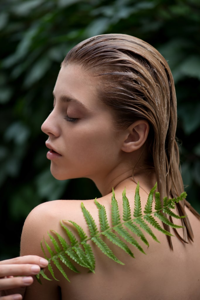 Beautiful young woman with perfect skin  front of plant tropical green leaves background with fern. Model applies a balm, shampoo or conditioner to her hair SPA, wellness, bodycare and skincare.